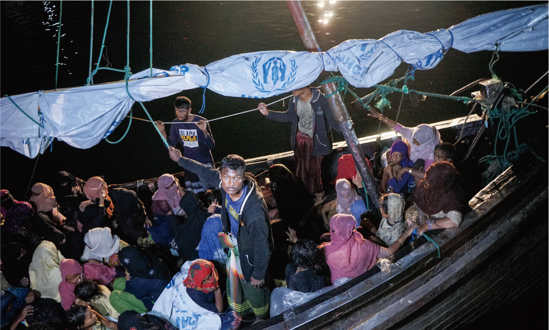 UNHCRのシートを掲げた木造船に大勢の避難民が乗り込んでいる夜の海上の様子